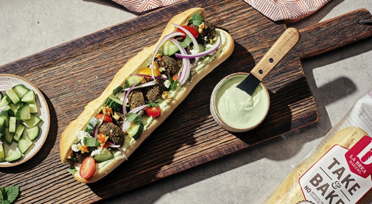 Fully prepared Falafel Sub with green chilli whipped feta and olive tapenade on wood cutting board with condiments