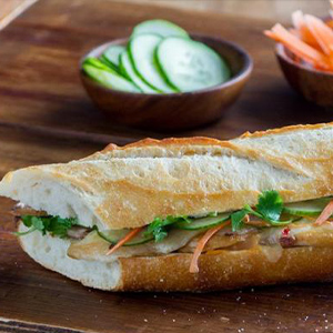 Banh Mi sandwich on a plate with cucumber slices and carrot chips in bowls on the side