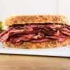 New York style pastrami on rye sandwich sitting on white serving board with pickles in the background