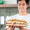 Smiling person holding a large sub sandwich filled with deli meats, shredded lettuce, cheese, and other toppings in a kitchen setting.