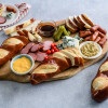 A charcuterie board featuring pretzel rolls, sliced sausages, blue cheese, pickles, cherry tomatoes, and various dipping sauces including a yellow cheese sauce and brown sauce.
