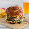 Gourmet burger with a glossy pretzel bun, melted cheese, crispy fried onions, jalapeños, and sauce, served on a wooden board with chips and a glass of beer.