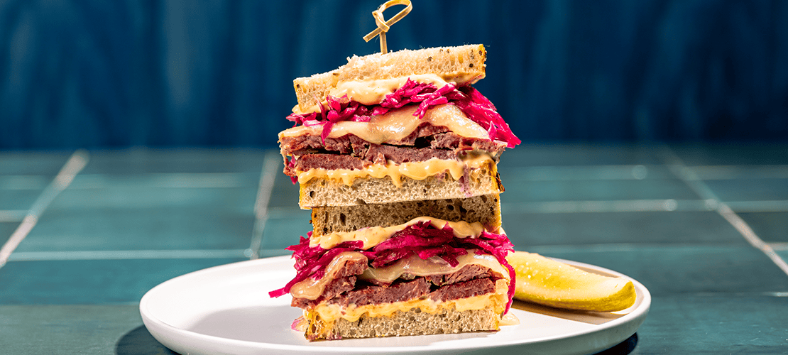 Reuben sandwich squares on New York Rye raisin bread stacked on a white plate with a pickle