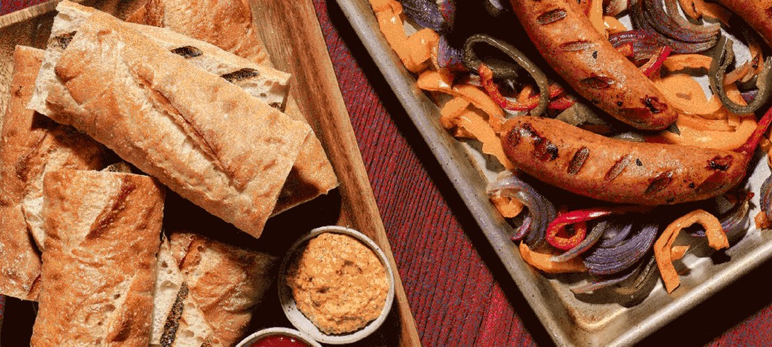 overhead shot of grilled sausages, peppers and onions with artisan bread in a metal serving tray laying on a picnic tabletop