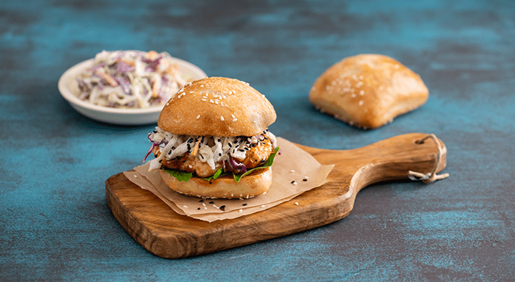 A gourmet burger on a wooden serving board, accompanied by a side of french fries in a small wire basket. The burger features a golden bun with sesame seeds, visible layers of meat patty, cheese, and fresh vegetables. The scene is set on a textured blue background, suggesting a rustic or casual dining presentation.