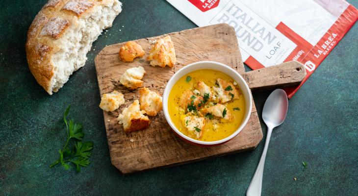 Bowl of Sweet Potato & Sage Soup with Croutons on a Wooden Board and a slice of Italian Round Loaf