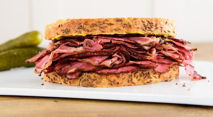 New York style pastrami on rye sandwich sitting on white serving board with pickles in the background