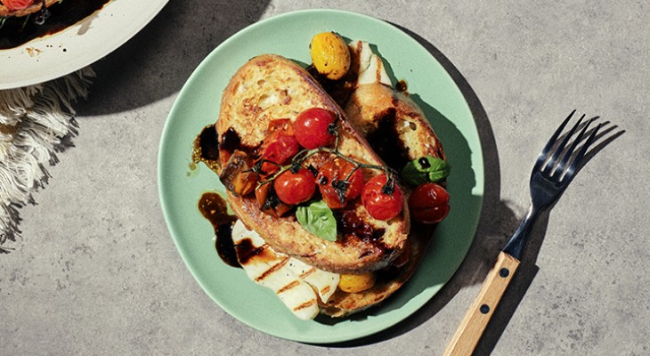 Two slices of rosemary round savory french toast topped with oasted tomatoes and halloumi on a green plate and concrete background