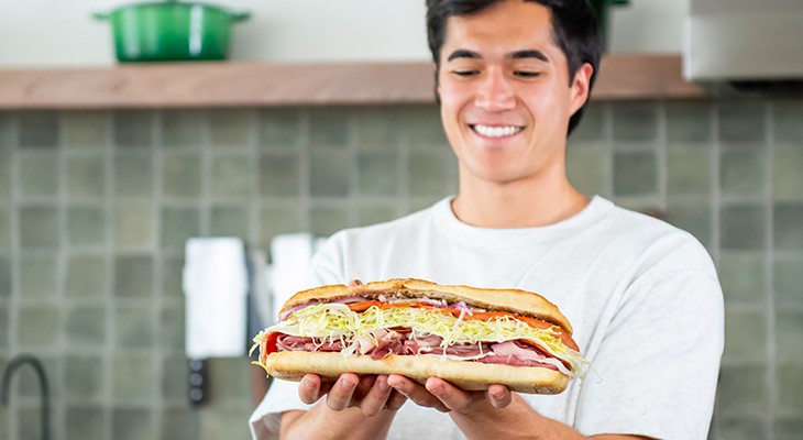 Smiling person holding a large sub sandwich filled with deli meats, shredded lettuce, cheese, and other toppings in a kitchen setting.