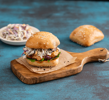 A gourmet burger on a wooden serving board, accompanied by a side of french fries in a small wire basket. The burger features a golden bun with sesame seeds, visible layers of meat patty, cheese, and fresh vegetables. The scene is set on a textured blue background, suggesting a rustic or casual dining presentation.