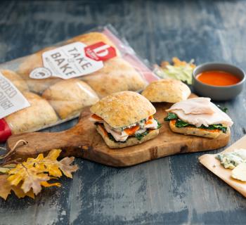 Buffalo Turkey Sliders on La Brea Bakery Seed Sourdough Rolls on a Cutting Board with Sides and a Bag of Rolls