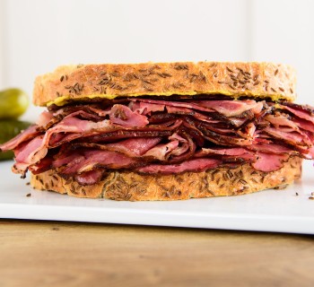 New York style pastrami on rye sandwich sitting on white serving board with pickles in the background