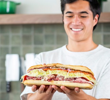 Smiling person holding a large sub sandwich filled with deli meats, shredded lettuce, cheese, and other toppings in a kitchen setting.